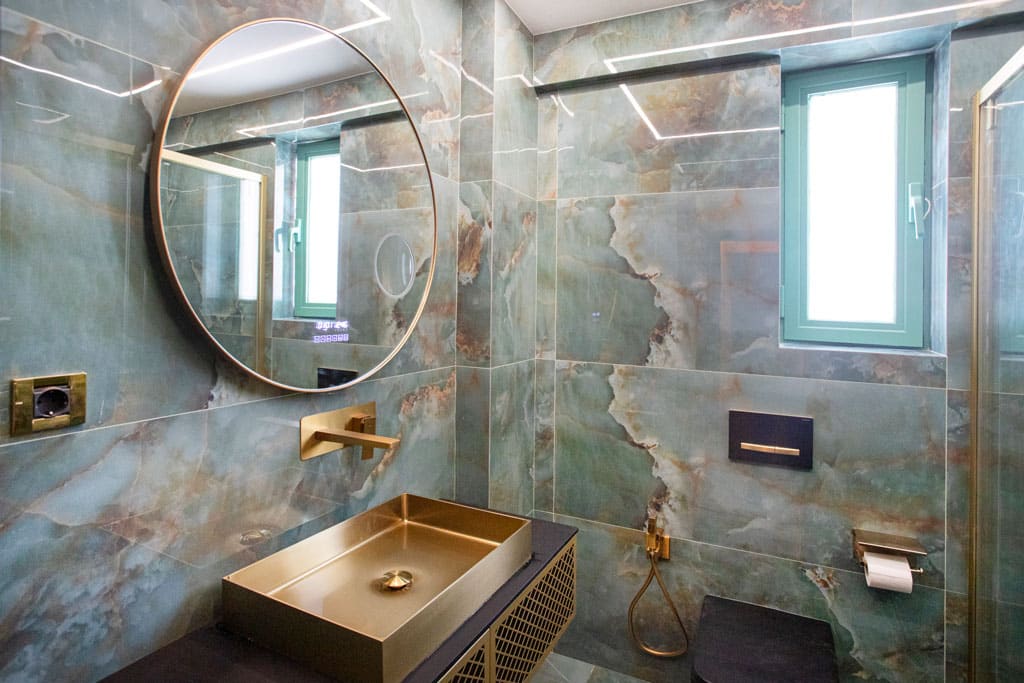 Ανακαίνιση μπάνιου στα Πατήσια με χρυσό νιπτήρα, στρογγυλό καθρέφτη, εντοιχισμένο καζανάκι και φυσικό φωτισμό από παράθυρο